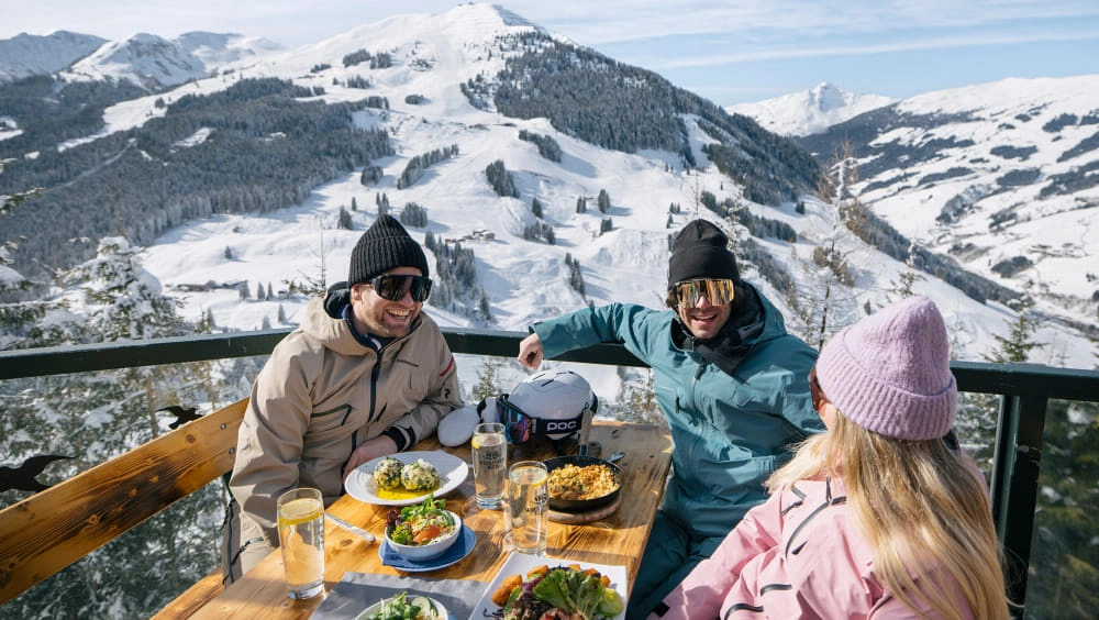 Three people enjoying food on a terrace with a winter mountain view.